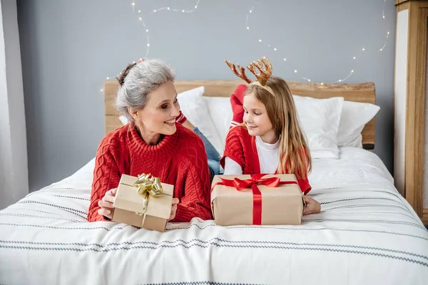Grandmother With Granddaughter In Santa Deer Horns on headt Are Lying On The Bed Holding Gifts And Smiling At Christmas. Family Having Fun During Christmas Festive