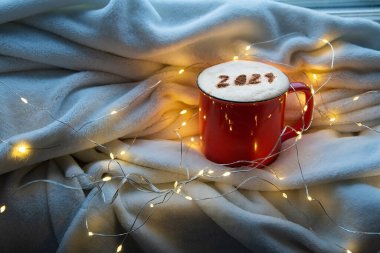 2021 numaralı kırmızı kahve fincanı sıcak ekose kapuçino ve Noel ışıkları arka planında köpüklü kapuçino. Mavi Gece Işığı. Mutlu yıllar 2021 yemek sanatı teması. Ayırıcı odak