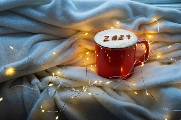 Xícara de café vermelho com o número 2021 na superfície espumosa de cappuccino no fundo acolhedor xadrez e luzes de Natal. Luz da noite azul. Feliz ano novo 2021 tema de arte alimentar. Foco selictivo — Fotografia de Stock