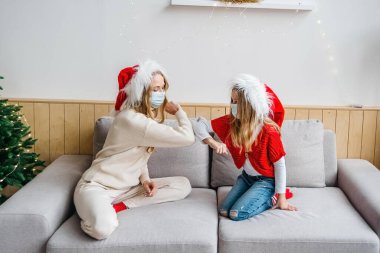 Dirseklerini sallıyor. Sosyal uzaklık ve sağlık hizmeti kavramı. Noel Baba şapkalı tatlı kız ve genç kadın koruyucu maskeler takıyor ve Noel süslemeli bir odada dirsekleriyle tokalaşıyorlar..