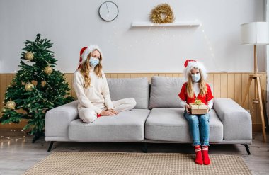 Noel 'i ve 2021 yılbaşını kutlarken sosyal mesafeler. Koruyucu maskeli iki kız koltukta oturuyorlar. Birbirlerinden 1 metre uzakta..