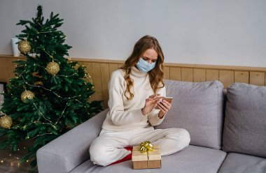 Oturma odasında akıllı telefondan yazı yazan genç bir kadın, Noel ağacı arka planında yüz maskesi takıyor. Covid-19 Noeli evde.
