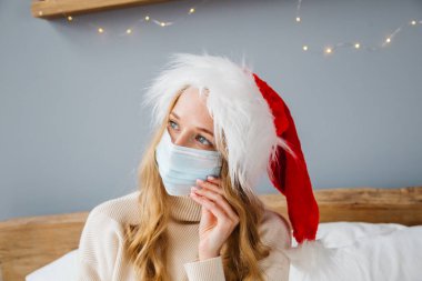 Sarı saçlı, Noel Baba şapkası takan ve maske takan tatlı, genç bir bayan Noel Baba..