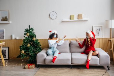 Noel 'i ve 2021 yılbaşını kutlarken yeni normal sosyal mesafeler. Ellerinde şampanya kadehleriyle koruyucu maskeli iki kız birbirlerinden uzakta koltukta oturuyorlar..