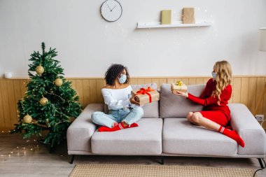 Koruyucu maskeli iki kız arkadaş kanepede oturuyor ve hediyelerini değiş tokuş ediyorlar. Covid-19 salgını sırasında Noel ve Yeni Yıl 2021 'i kutlarken sosyal uzaklık.