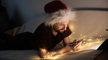Yakışıklı genç adam kanepeye uzanmış Noel Baba şapkası takıyor, telefonunu kullanıyor ve karanlık noel süslemeli odasında gülümsüyor. Noel ve Yeni Yıl kutlamalarında vakit geçirmek çok hoş..