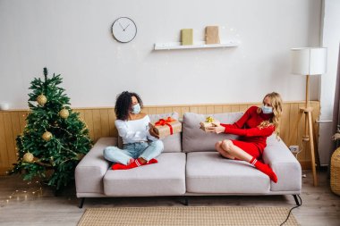Koruyucu maskeli iki kız arkadaş kanepede oturuyor ve hediyelerini değiş tokuş ediyorlar. Covid-19 salgını sırasında Noel ve Yeni Yıl 2021 'i kutlarken sosyal uzaklık.