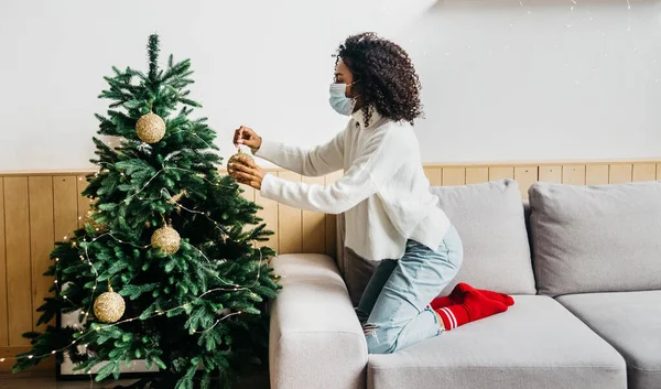 Giovane donna afroamericana indossando maschera protettiva e decorare l'albero di Natale a casa. Distanziamento sociale e prevenzione del coronavirus durante i festeggiamenti di Natale e Capodanno 2021 — Foto Stock
