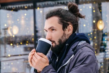 Açık havada sıcak kahve içen çekici sakallı genç adamın portresi. Bir adam kışın kendini sıcak kahveyle ısıtır. Servis dışı.