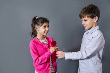 Gri arka planda kırmızı kalpli lolipop taşıyan mutlu çocuklar. Çocuk, sevgililer günü lolipopunu kıza vermek istiyor. Sevgililer Günü