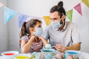 Yeni normal yaşam tarzı konsepti. Tatlı küçük kız babası koruyucu maskeler takıyor. Paskalya hazırlarken eğleniyor. Coronavirus salgını sırasında evde yumurta boyuyor..