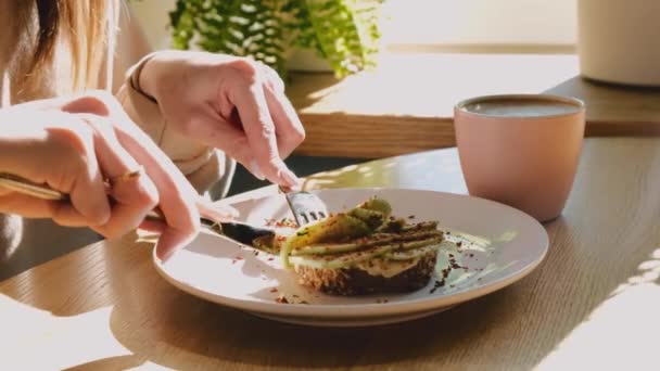 Close up de mãos femininas cortando uma deliciosa torrada com húmus, abacate com um garfo e faca. Pequeno-almoço saboroso e saudável. — Vídeo de Stock
