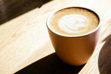 Bir kupa kapuçino ahşap masanın üzerinde, pencerenin yanındaki kafede, koyu gölgeli güneş ışığı altında..