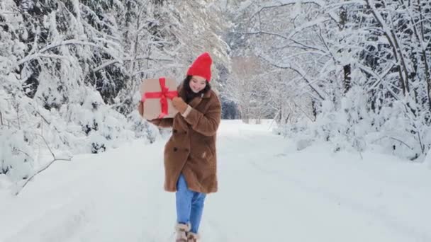 Una joven con un abrigo de piel de peluche marrón y un sombrero rojo sostiene una gran caja de regalo con una cinta roja y camina en el bosque de invierno de nieve. Estilo de vida festivo y concepto de compras de regalos de Navidad. — Vídeo de stock