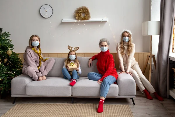 Coronavirus pandemia distancia Navidad y Año Nuevo concepto. Familia femenina con máscaras protectoras y sentada en la sala de estar de Navidad. Imagen De Stock