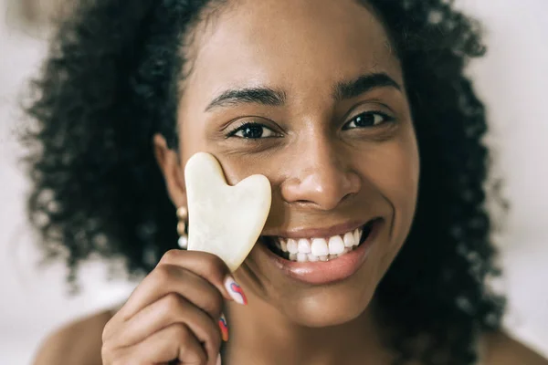 Primer plano retrato de sonriente hermosa mujer multiétnica usando facial gua sha jade, mirando a la cámara. Concepto de autocuidado Fotos De Stock Sin Royalties Gratis