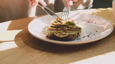 Lezzetli sağlıklı kahvaltı konsepti. Kadın ellerinin enfes bir tostu humusla, avokadoyu çatal ve bıçakla dilimlemesi. 4K, ağır çekim.