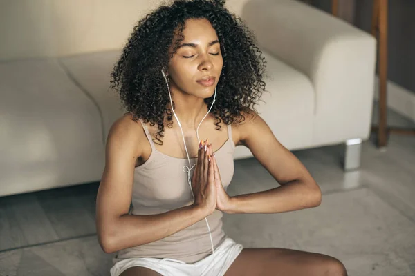Mentális egészség koncepció. Fiatal fekete nő gyakorolja meditáció otthon, ül Easy Seat pózban, a szeme csukva, hallgat audio meditáció fülhallgatóval a telefonnal Stock Kép