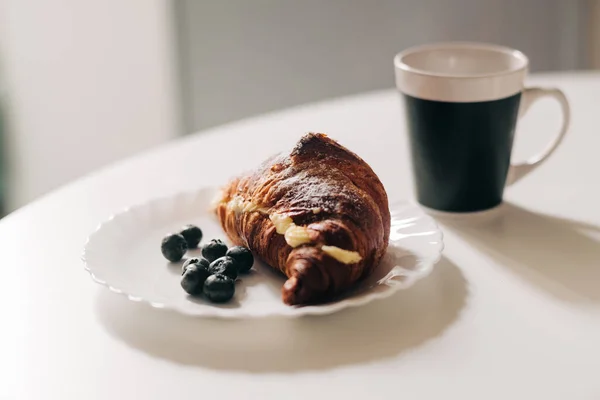 Croissant mit Sahne und Blaubeere auf einem weißen Teller, Tasse Kaffee im Hintergrund. Kontinentales Frühstückskonzept — Stockfoto