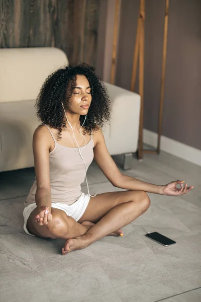 精神保健ライフスタイルの概念の傾向。自宅で瞑想を練習する若い女性は、イージーシートのポーズに座って、オーディオ瞑想を聞く。垂直写真 ロイヤリティフリーのストック画像