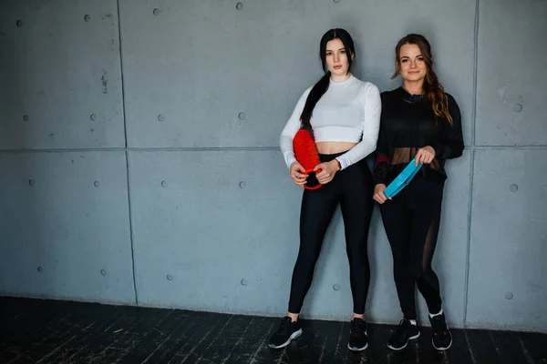 Dos atletas femeninas están de pie contra el fondo de una pared gris y sosteniendo rollo de masaje y banda de goma de entrenamiento —  Fotos de Stock