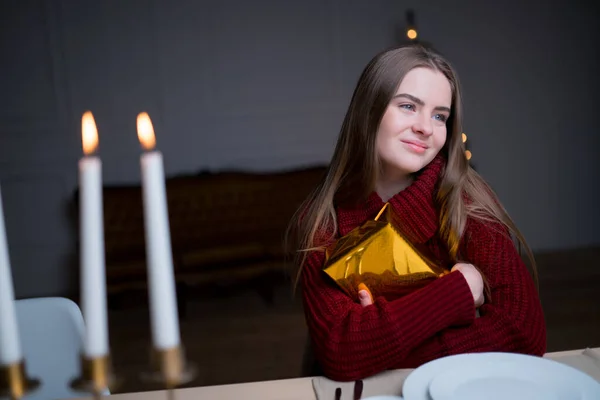 Menina caucasiana sentada em uma mesa festiva com velas de Natal, abraçando uma caixa de presente e se alegrando — Fotografia de Stock