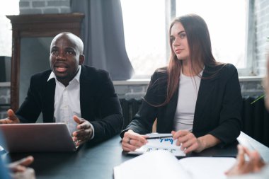 İki iş ortağı, bir Afrikalı Amerikalı erkek ve bir Avrupalı kadın ortaklarla bir ofis masasında oturuyorlar. Not Defteri, Grafikler, Kâr Büyütme İstatistikleri