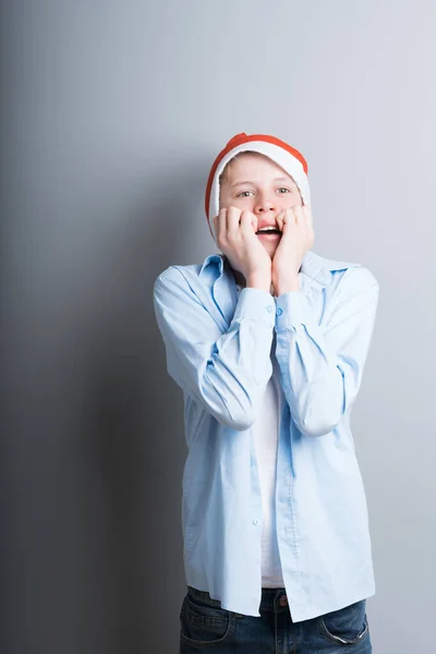 Um menino de 12 a 15 anos de idade em um chapéu de Natal vermelho fica em um fundo cinza em antecipação ao ano novo. Um jovem se alegra em antecipação ao Natal — Fotografia de Stock