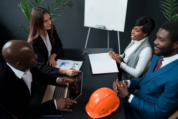 A group of diverse colleagues are sitting at a table and discussing business ideas in a loft office. Partners or office workers working together. Orange construction hard hat on office desk