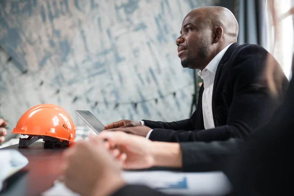 Мужчина африканский американский директор строительной компании проводит встречу с коллегами, садится за стол с документами и оранжевым строительным шлемом — стоковое фото