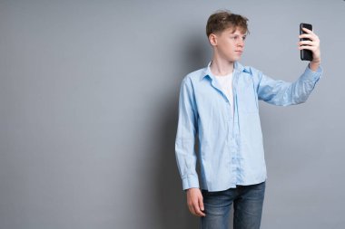 Young boy in a blue shirt and blue jeans makes a selfie on the phone, stands on a gray background