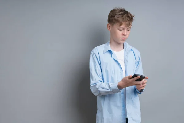 Um adolescente de camisa azul e uma camiseta branca toca o telefone em um fundo cinza. Adolescente problemático, questões sociais, jogador, vício em telefone — Fotografia de Stock