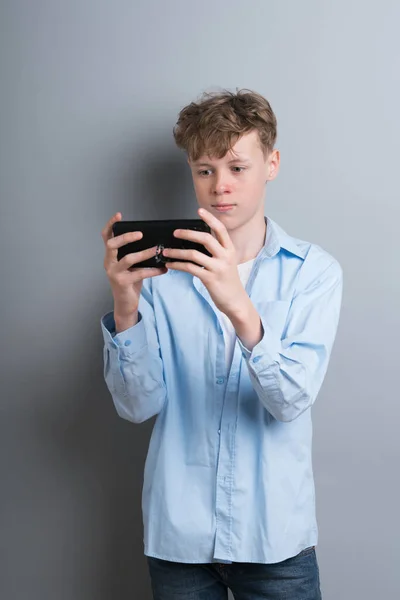 Nastolatek w niebieskiej koszuli i białej koszulce gra telefon na szarym tle. Dotknięty nastolatek, problemy społeczne, gracz, uzależnienie od telefonów — Zdjęcie stockowe