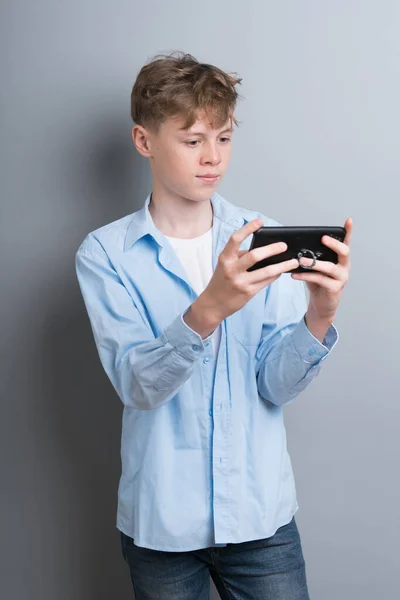 Un adolescente con una camisa azul y una camiseta blanca toca el teléfono sobre un fondo gris. Adolescente con problemas, problemas sociales, jugador, adicción al teléfono — Foto de Stock