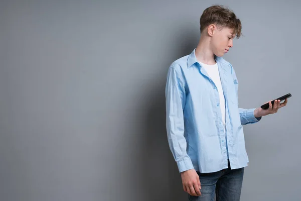 A teenager boy in a blue shirt and a white T-shirt plays the phone on a gray background. Troubled teenager, social issues, gamer, phone addiction