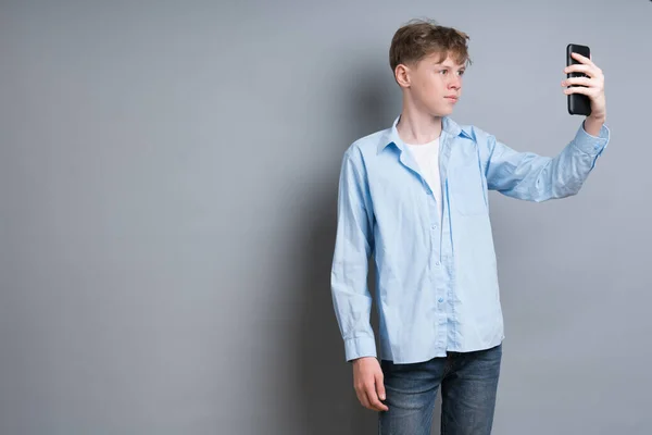 Young boy in a blue shirt and blue jeans makes a selfie on the phone, stands on a gray background — Fotografia de Stock