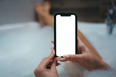 Köpük banyosunda oturan bir kadının elindeki beyaz bir telefon ekranının yakın çekimi. Mesaj için yer var. Rahatlama ve rahatlama