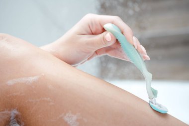 Yan görüş. Dişi bir bacağa yakın çekim. Bir kadın köpük banyosunda yatarken bacaklarını jiletle tıraş eder.