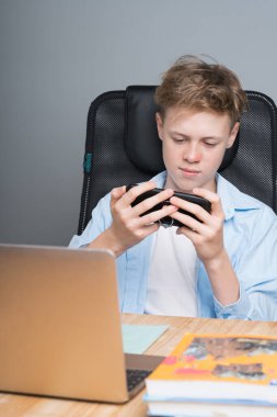 Beyaz çocuk bir masada oturuyor, telefonda oynuyor, online bir derste oturuyor. Evde eğitim, okul çocuğu, salgın dersler.