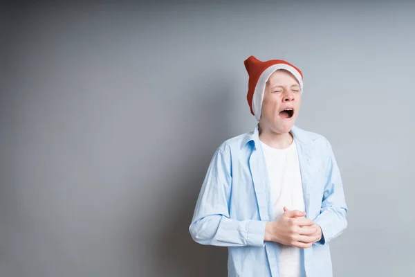 Menino 13-15 anos de idade em uma camisa azul, um chapéu de Natal vermelho boceja em um fundo cinza. À espera da noite de Natal, conceito de férias — Fotografia de Stock