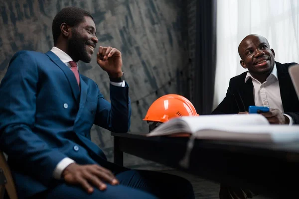 Two African American men listen to a speaker at a business training in the office. Workers sit at a table with a folder for documents and a construction helmet
