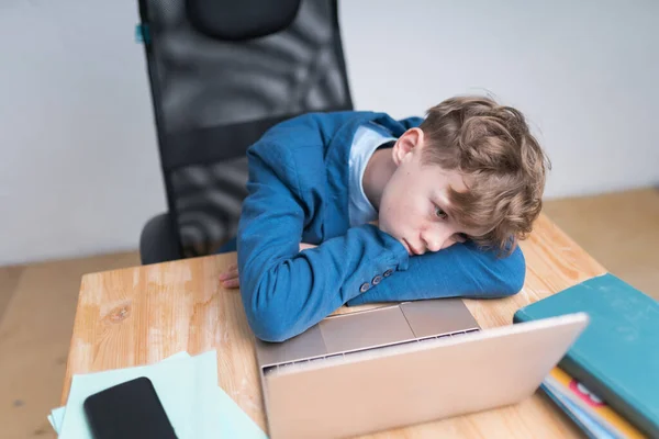 Menino loiro de uniforme escolar azul está entediado durante suas aulas online. Ele está quase dormindo, deitado com a bochecha no braço, olhando para a tela sem qualquer interesse — Fotografia de Stock