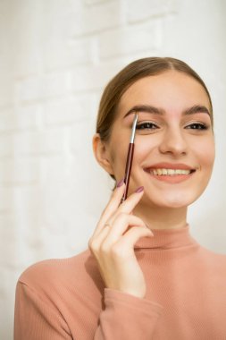 Kaşlarına kozmetik ürünle fırça süren bir kız. Güzel tatlı kız gülümser ve kaş makyajı yapar.