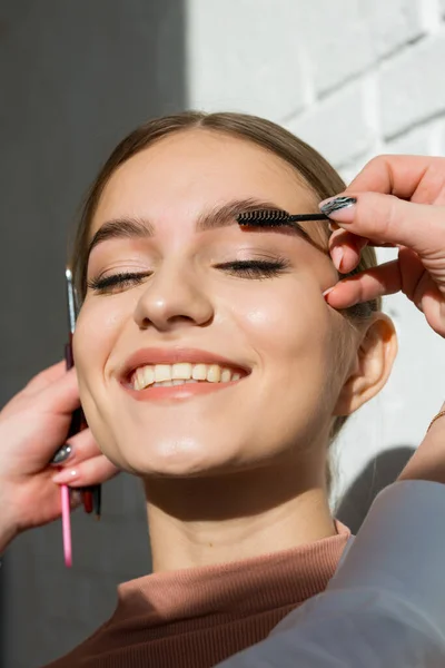 Beautiful attractive girl with natural makeup smiles in the sunlight. Makeup artist combing eyebrows with a brush — Stock Photo, Image