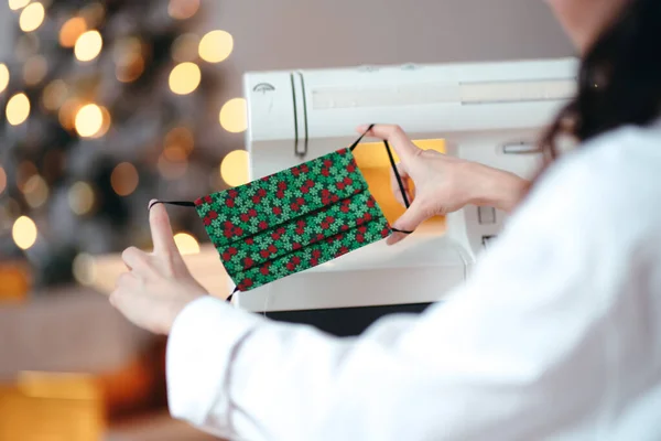 Close-up. Máscara médica de Natal em mãos femininas no fundo de uma máquina de costura e uma árvore de Natal — Fotografia de Stock