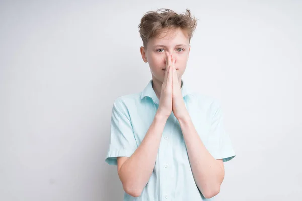 Um menino caucasiano em um fundo branco segurando as palmas das mãos juntas em uma pose de oração. O cabelo dele está confuso, os olhos tranquilos e calmos. — Fotografia de Stock