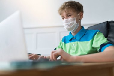 Tek kullanımlık maske takan yakışıklı Avrupalı çocuk karantina sırasında dizüstü bilgisayarında ödev yapıyor. Çevrimiçi çalışma kavramı