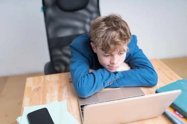 Menino caucasiano de uniforme escolar está entediado durante as aulas online. Está deitado com a bochecha no braço, desinteressado. — Fotografia de Stock