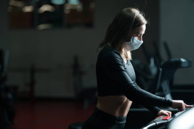 Koronavirüs salgını sırasında spor salonunda koşu yaparken koruyucu yüz maskesi takan bayan sporcu. Koşu bandında ısınma turu.