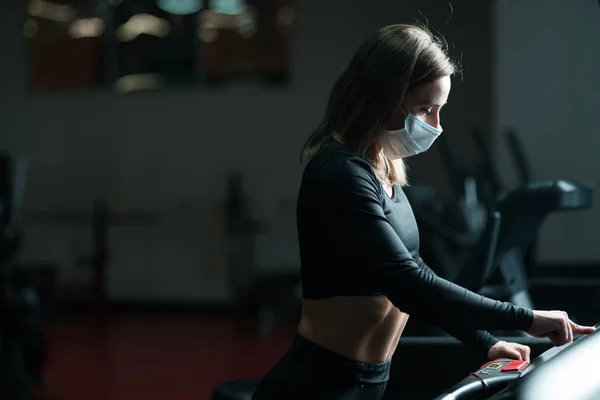 Atleta femenina corriendo en pista mientras usa mascarilla protectora en un gimnasio durante la epidemia de coronavirus. calentamiento en una cinta de correr —  Fotos de Stock